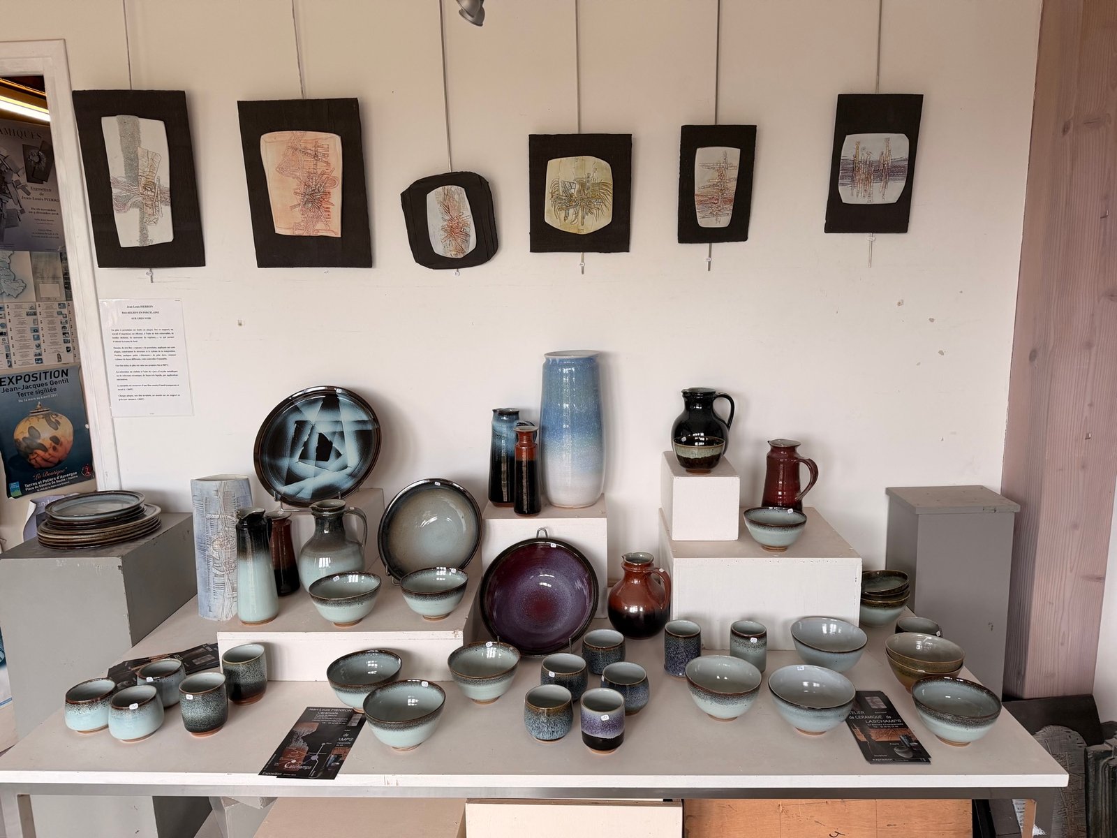 Salle d’exposition : six plaques de porcelaine au mur, vases, bols et pichets sur la grande table