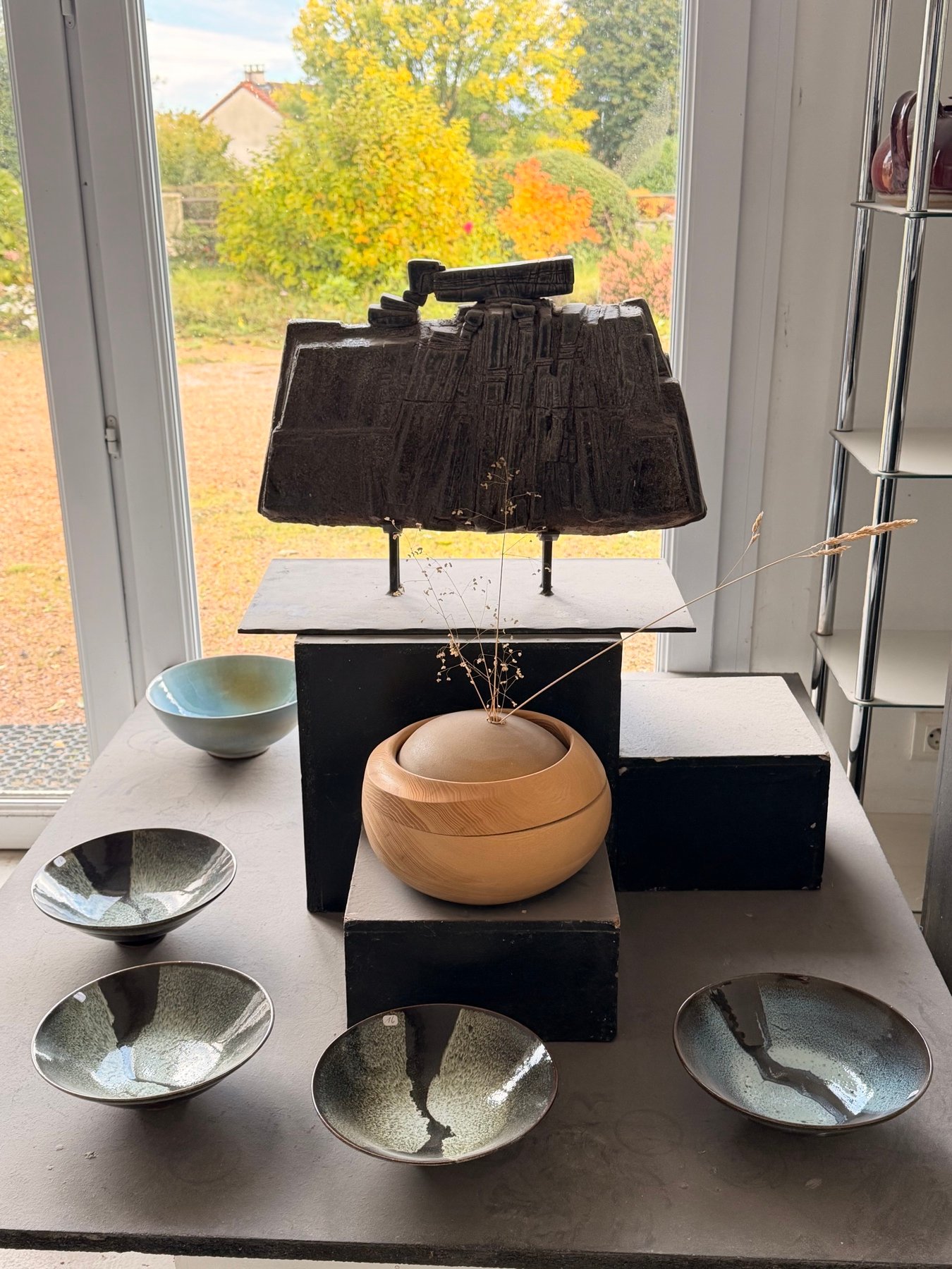 Une sculpture noire sur socle, un pot en bois et des coupes en céramique posés devant une fenêtre qui s’ouvre sur le jardin en automne