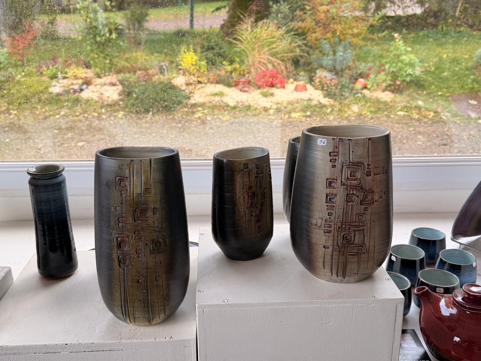 Trois vases en céramique sombre aux motifs carrés incisés, devant une fenêtre sur le jardin