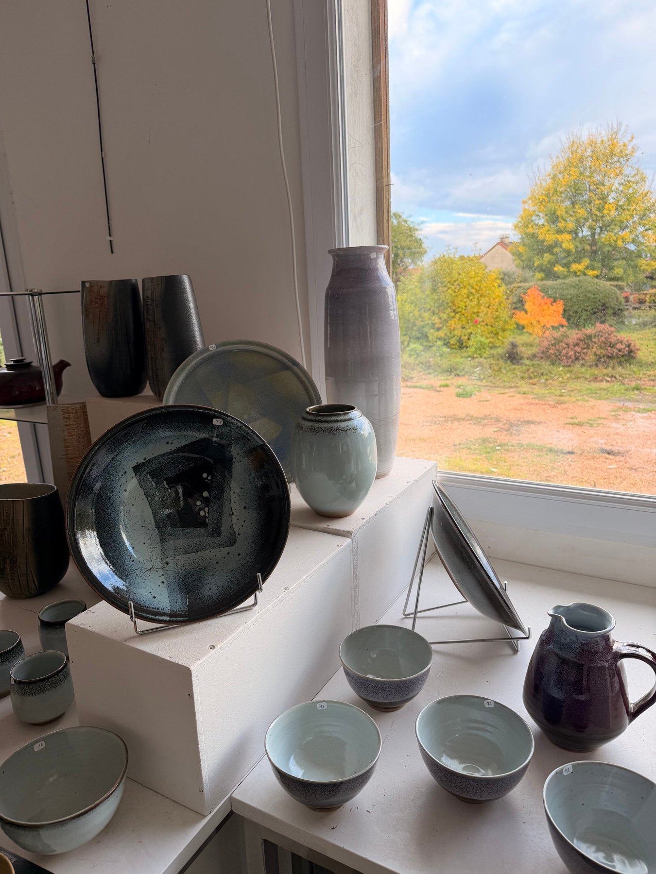 Vitrine près de la fenêtre : grande assiette bleu-noir, vases, bols et pichet